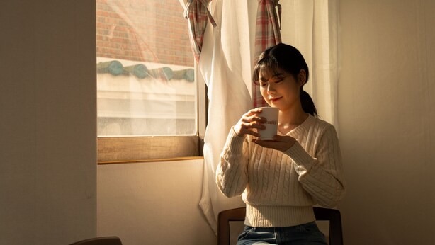 A person sitting by a window with curtains, holding a cup. - Oak&Co (Seoul)