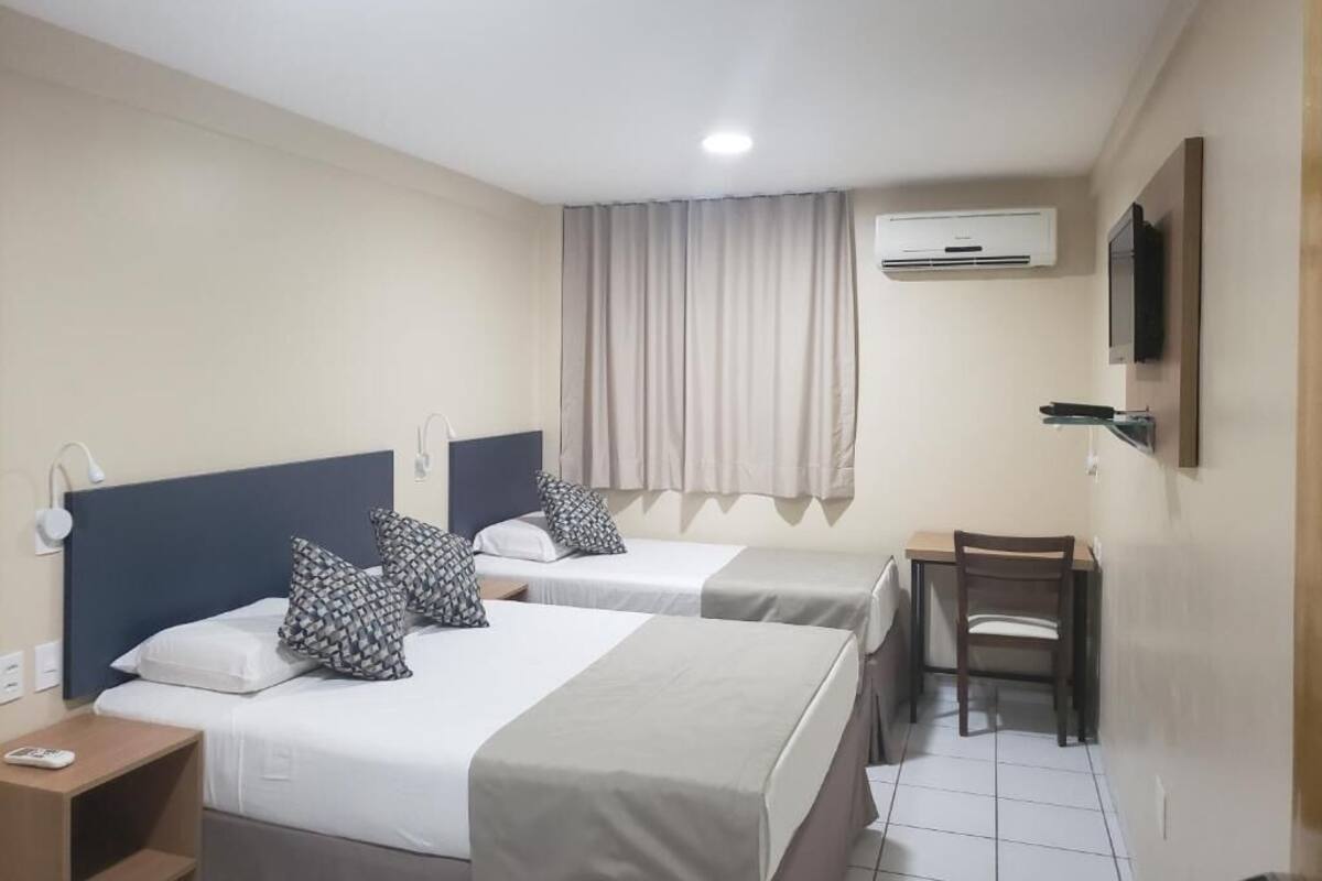 A hotel room with two beds, a desk, a chair, an air conditioning unit, and a television mounted on the wall.