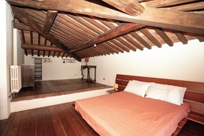 A bedroom with a wooden ceiling, a bed with white pillows, a radiator, and a table with a lamp.