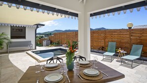 Un coin repas avec une table en bois dressée pour quatre personnes, entourée de chaises, d’une petite piscine et d’une clôture en bois.