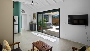 Un vaste espace de vie avec une grande porte-fenêtre coulissante donnant sur une terrasse, un téléviseur à écran plat fixé au mur et une table en bois sur laquelle se trouve une télécommande.