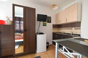 Ein kleines Zimmer mit einem Bett, einem kleinen Kühlschrank, einem Fernseher, einer Küchenzeile und einem Glastisch.