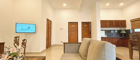 A living room with a sofa, a wooden coffee table, and a TV mounted on the wall. There are two wooden doors and a kitchen area visible in the background.