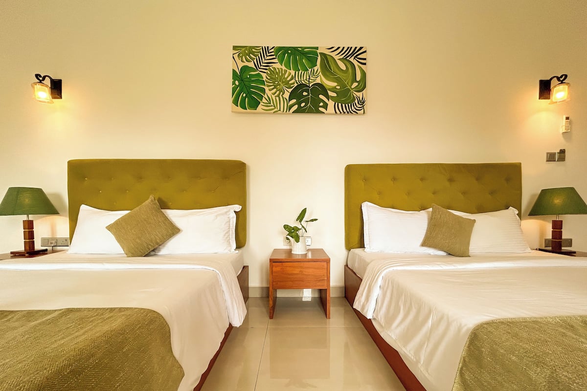 Ein Hotelzimmer mit zwei Betten, jeweils mit einem grünen Kopfteil, weißer Bettwäsche und einem kleinen Nachttisch mit einer Lampe. Über den Betten ist ein an der Wand befestigtes Kunstwerk mit tropischen Blättern zu sehen.