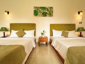 A hotel room with two beds, each with a green headboard, white bedding, and a small bedside table with a lamp. A wall-mounted artwork with tropical leaves is visible above the beds.