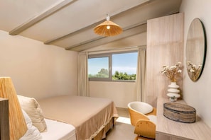 A bedroom with a bed, a chair, a vanity with a mirror, a vase with flowers, and a view of greenery through the window.