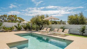 A rectangular swimming pool with clear water, surrounded by a white fence, and equipped with lounge chairs and a shaded umbrella.