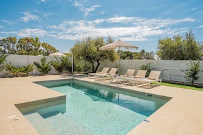 A rectangular swimming pool with clear water, surrounded by a white fence, and equipped with lounge chairs and a shaded umbrella.