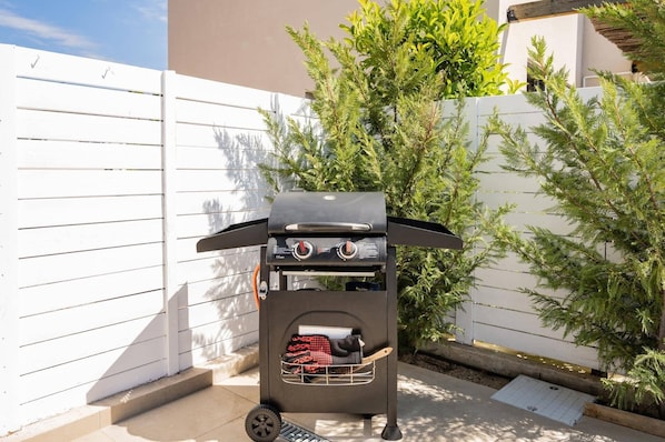 A portable barbecue grill with a side compartment for food storage, positioned on a patio with a white fence and green foliage in the background.
