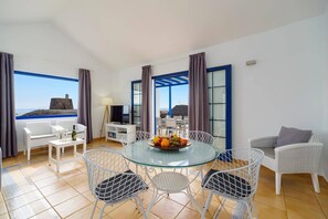 A room with a glass table, white wicker chairs, a TV, and a view of the sea.