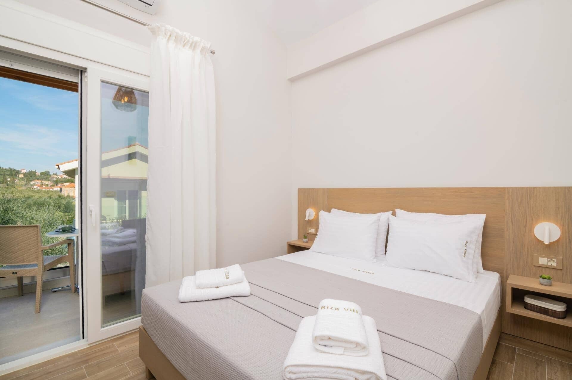 A hotel room with a bed, a nightstand, a window with a view of the outside, and a sliding glass door leading to an outdoor area.
