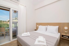 A hotel room with a bed, a nightstand, a window with a view of the outside, and a sliding glass door leading to an outdoor area.