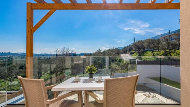 En balkon med et bord, stole, en potteplante og udsigt til en vej og grønt område.