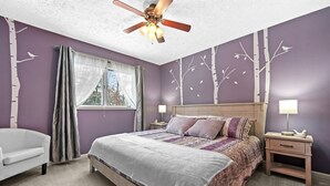Une chambre à coucher comprenant un lit, des tables de chevet, un ventilateur de plafond, une fenêtre avec des rideaux et des autocollants décoratifs représentant des arbres sur les murs.