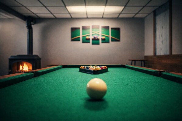A billiards table with a cue ball in the center, surrounded by a green felt surface. There is a set of billiard balls and a fireplace on the left side. - Fall Escape | Hot Tub + Games | Near Bass Lake (Traverse City)