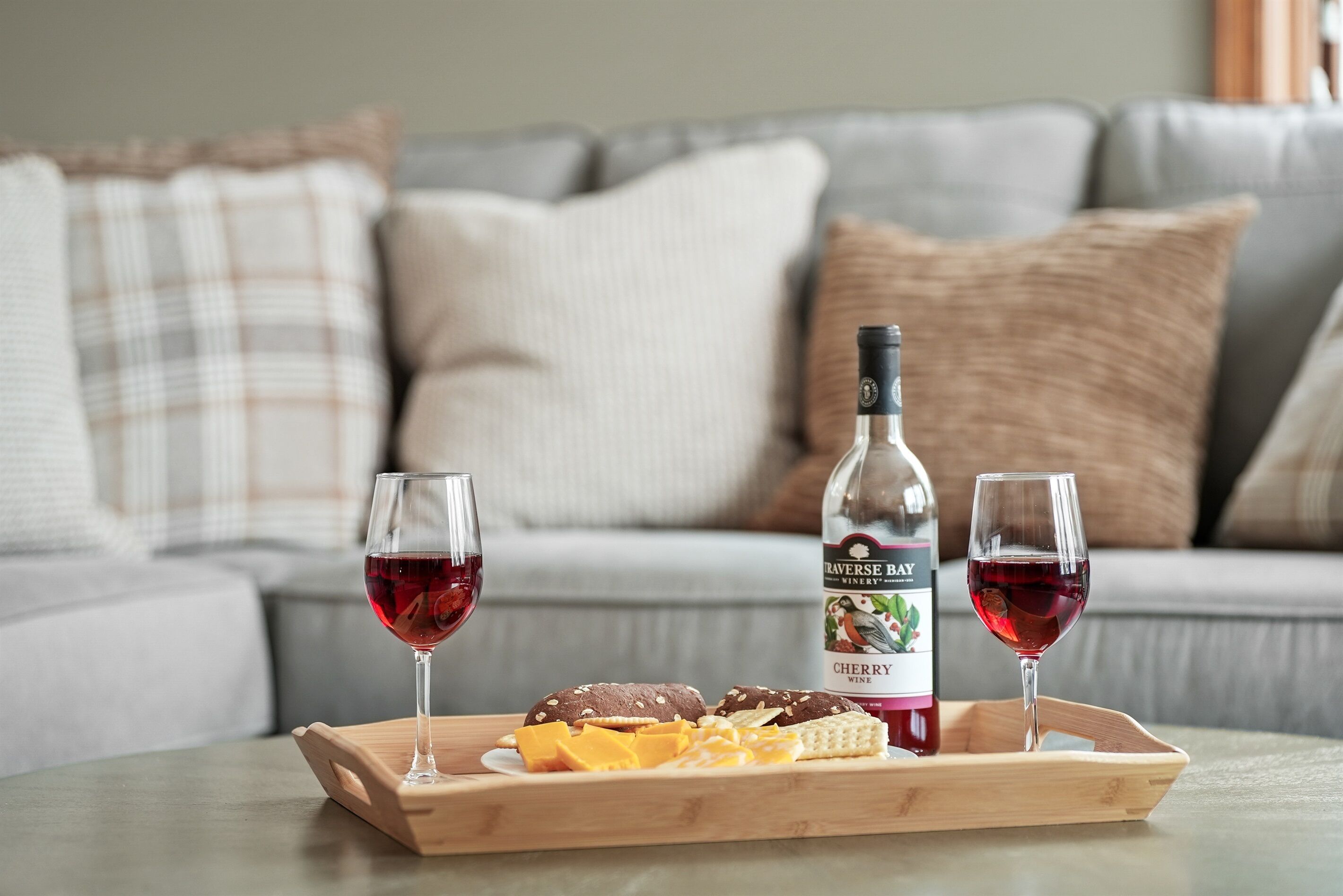 Un plateau en bois avec deux verres de vin rouge, une bouteille de vin et une sélection de fromages et de crackers, le tout posé sur une table devant un canapé gris agrémenté de coussins.
