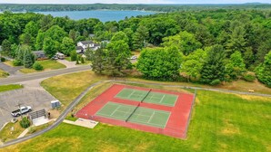 Eine Vogelperspektive auf einen Tennisplatzkomplex mit zwei Plätzen, umgeben von dichtem Wald und einem See im Hintergrund.