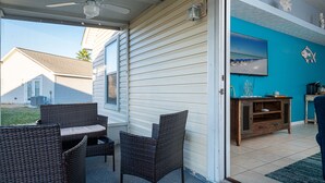Une terrasse couverte, meublée de mobilier en osier, dotée d’un ventilateur de plafond et offrant une vue sur une maison.