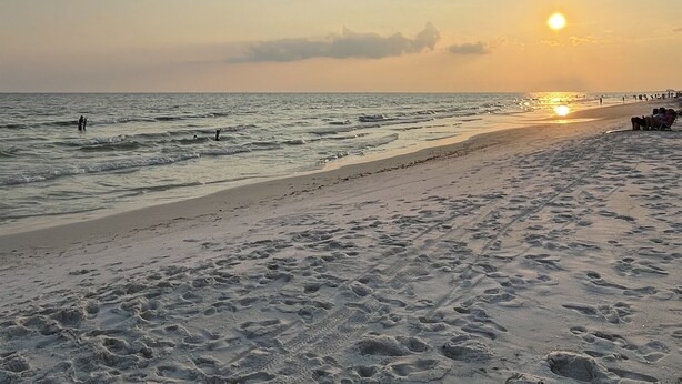 Une plage où l’on voit des empreintes de pas dans le sable, un coucher de soleil en arrière-plan et une jetée animée par la présence de personnes.
