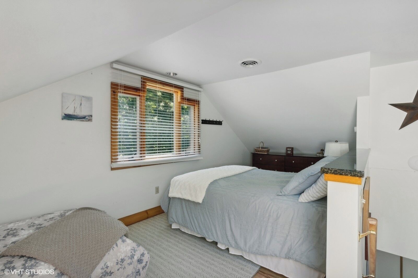 Een slaapkamer met een schuine kap, een bed met een blauwe dekbedovertrek, een dressoir, een raam met jaloezieën en een sterren decoratie aan de muur.