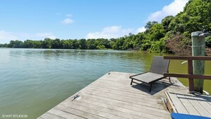 Un muelle de madera con una silla y una sombrilla, con vistas a un lago tranquilo y abundante vegetación en la orilla.