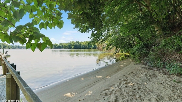 Una playa arenosa con un camino de madera que lleva a un lago tranquilo, rodeado de abundante vegetación verde.