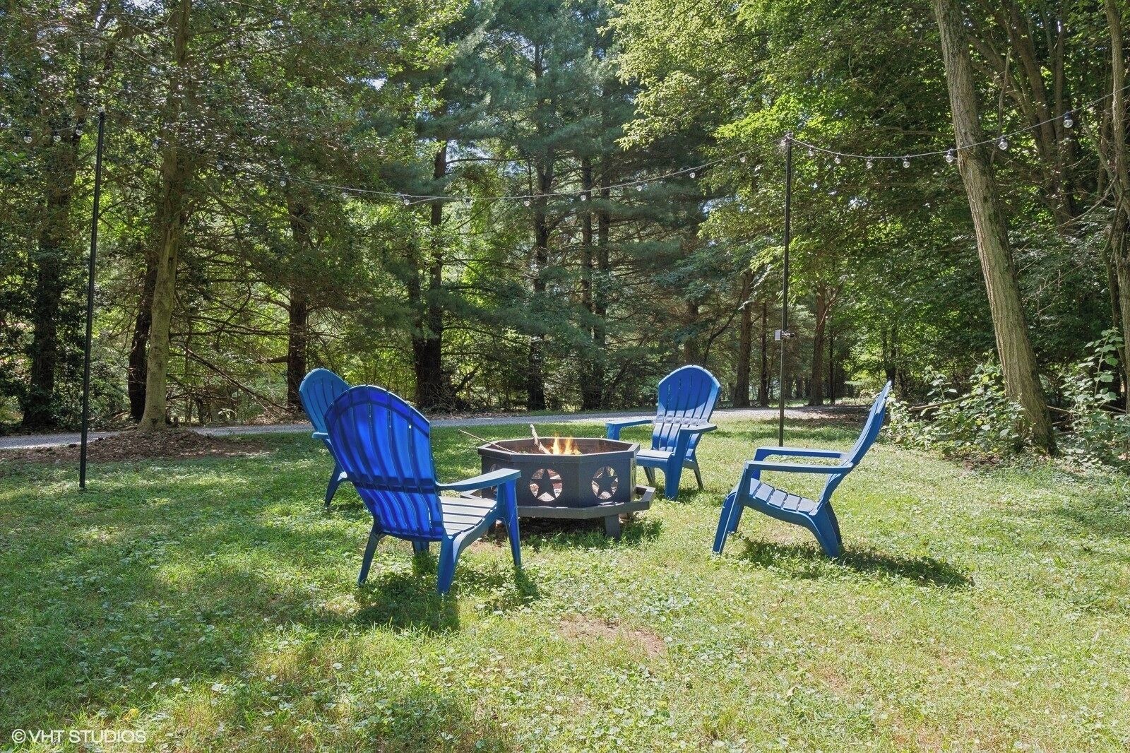 A forested area with a fire pit and blue Adirondack chairs.