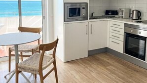 Eine moderne Küche mit weißen Schränken, einer Mikrowelle, einem Wasserkocher und einem Backofen. Ein runder Tisch mit Stühlen bietet Blick auf den Ozean durch eine Glastür.