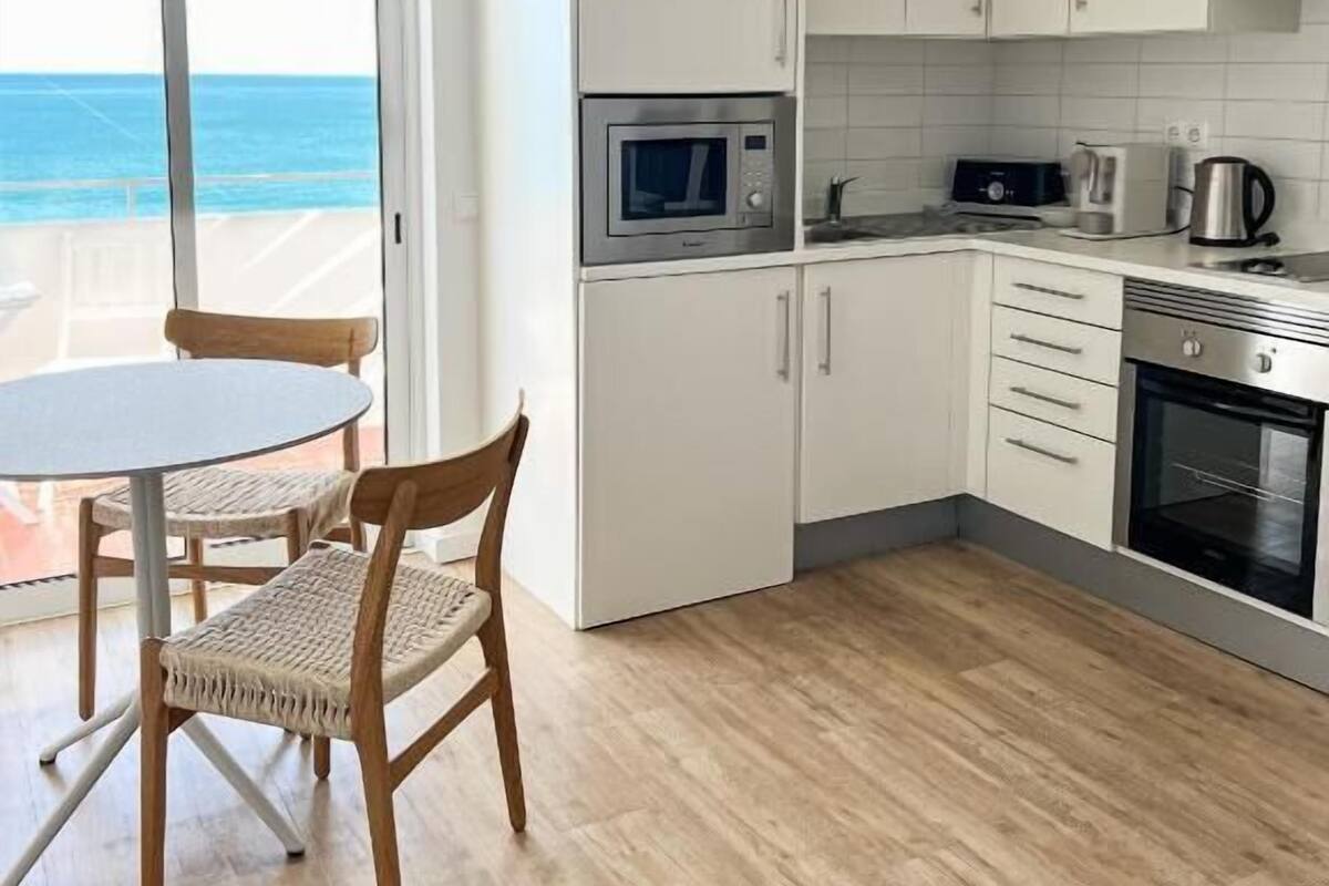 A modern kitchen with white cabinets, a microwave, a kettle, and an oven. There's a round table with chairs and a view of the ocean through a glass door.