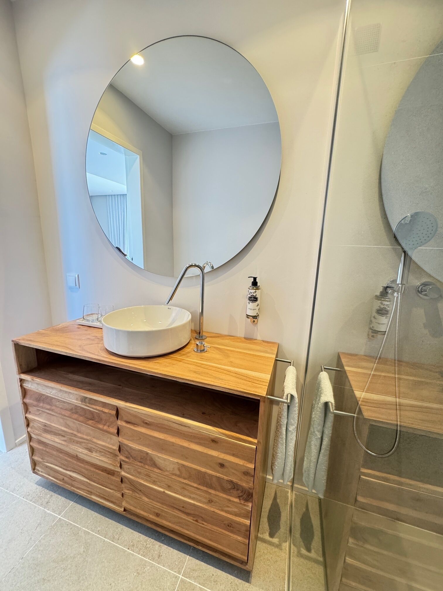 A bathroom with a round mirror, a sink, a wooden vanity, and a glass shower enclosure.