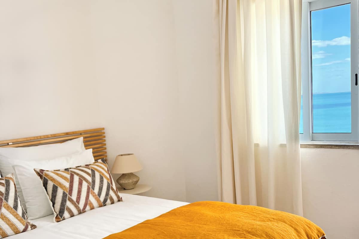 A neatly made bed with a mustard-colored blanket, a wooden headboard, and patterned pillows. A bedside lamp is on a small table beside the bed.
