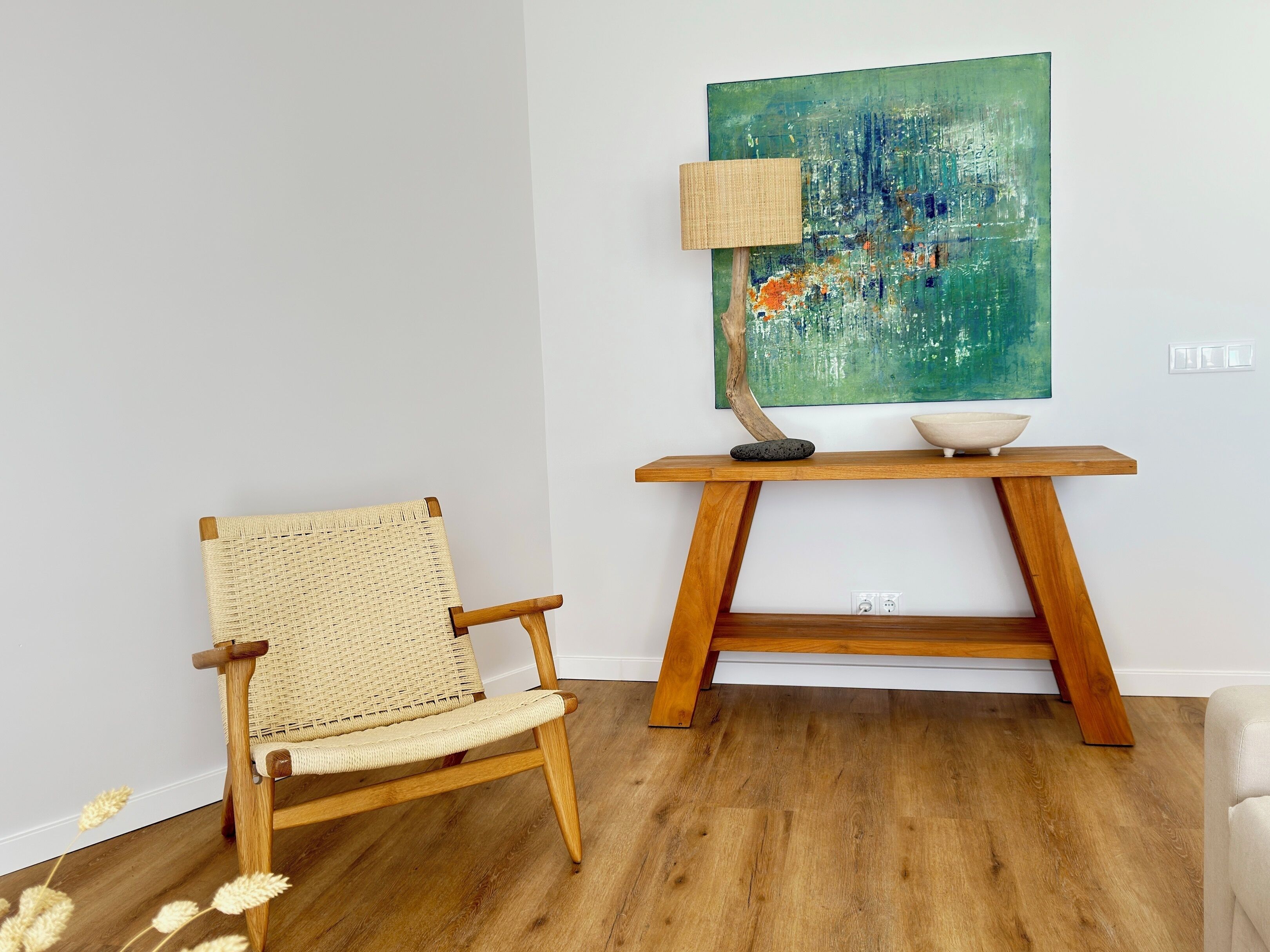 Una habitación con una silla de madera, una mesa con un cuenco y una lámpara, y un cuadro en la pared.
