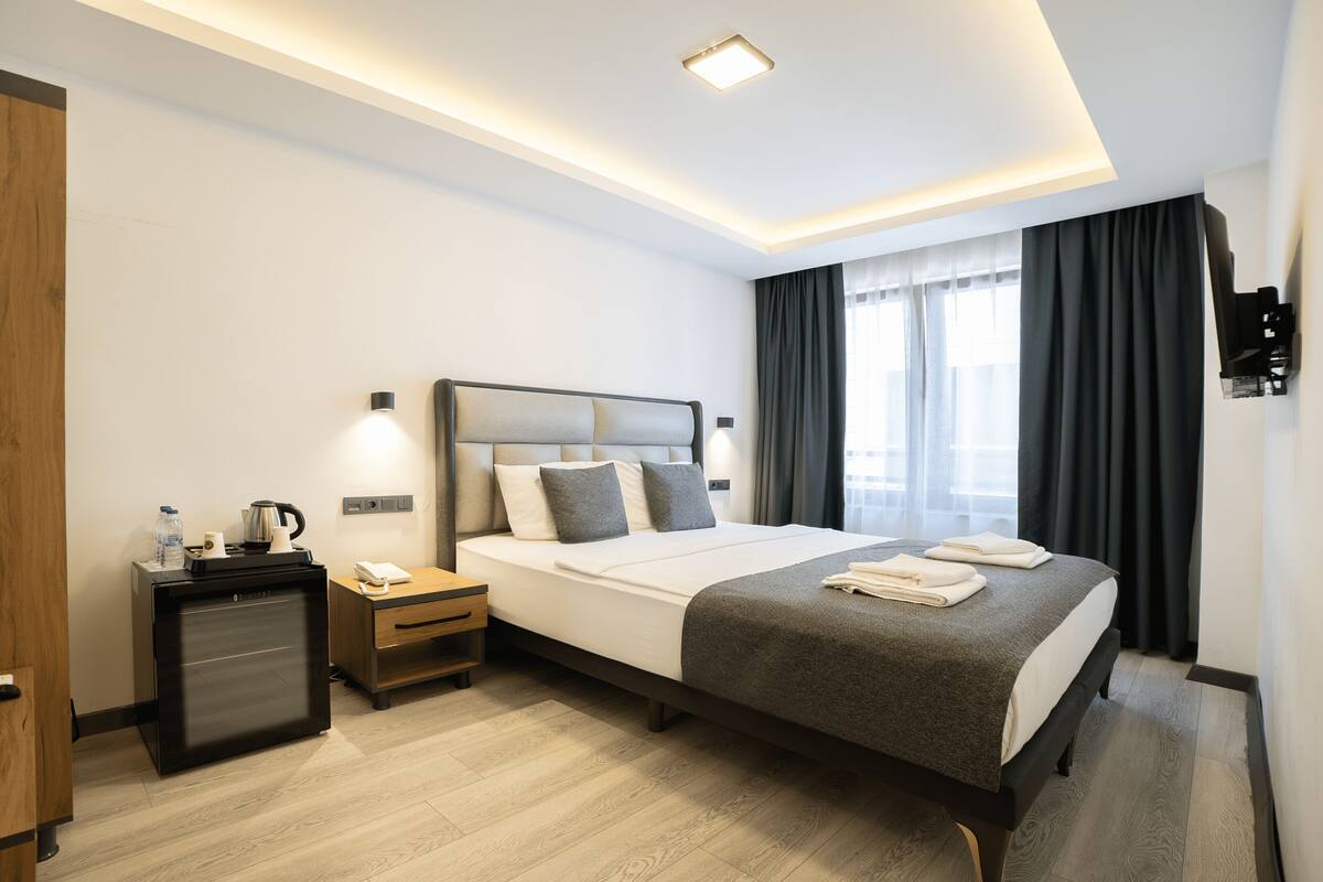 A modern hotel room with a neatly made bed, a bedside table with a lamp, a small desk with a kettle, and a wardrobe.