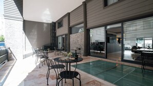 Un área de patio al aire libre con varias mesas y sillas, una partición de vidrio y un mostrador de recepción visible a través de las puertas de cristal.