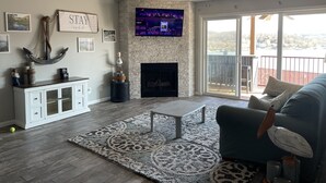 A spacious living room with a fireplace, a mounted TV, and a balcony with a view. - Lakeview Luxury: Stunning 2 Bed, 2 Bath Condo with Breathtaking Lake Views (Camdenton)