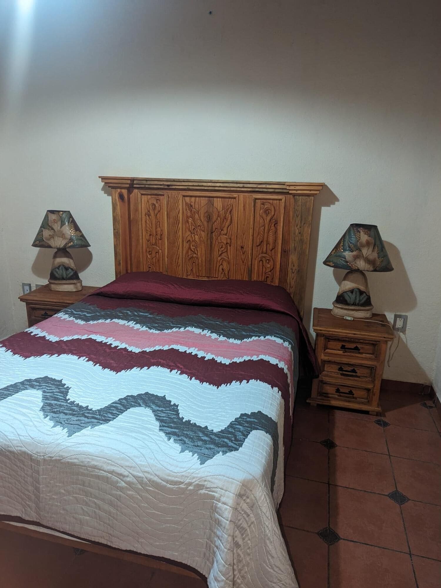 Un dormitorio con cabecera de madera, colcha estampada, dos mesitas de noche con lámparas y suelo de baldosas.
