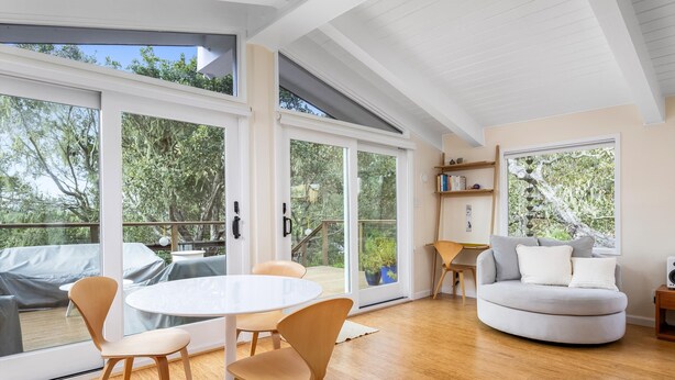 A modern living room with a round coffee table, two chairs, a sofa, and a bookshelf. Large windows provide views of the outdoors. - Tranquil Treehouse - EV Charger (Cambria)