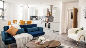 Una sala de estar moderna con un sofá azul, un sillón blanco y una mesa de centro de madera. Se ve una cocina con gabinetes blancos y una estufa al fondo.