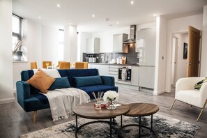 A modern living room with a blue sofa, a coffee table, and a kitchen in the background. - Cityscape Haven - Leicester's Premier Retreat (Leicester)