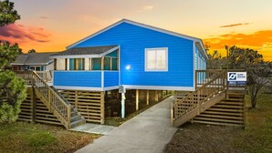 Une maison bleue avec un porche et un escalier en bois, entourée d’arbres, avec un coucher de soleil en arrière-plan.
