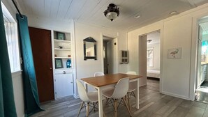 Een eetgedeelte met een houten tafel en witte stoelen, een keuken met witte kasten, en een gang die naar een andere kamer leidt.