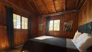 A wooden cabin bedroom with a bed, a painting on the wall, and a window with curtains. - 2 BR, walk to beach, volleyball (Sanilac Township)