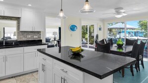 Cocina moderna con gabinetes blancos, encimera negra y una isla central con fruta. Hay luces colgantes y un ventilador de techo.