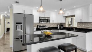 Una cocina moderna con gabinetes blancos, electrodomésticos de acero inoxidable, una isla central con taburetes e iluminación colgante.