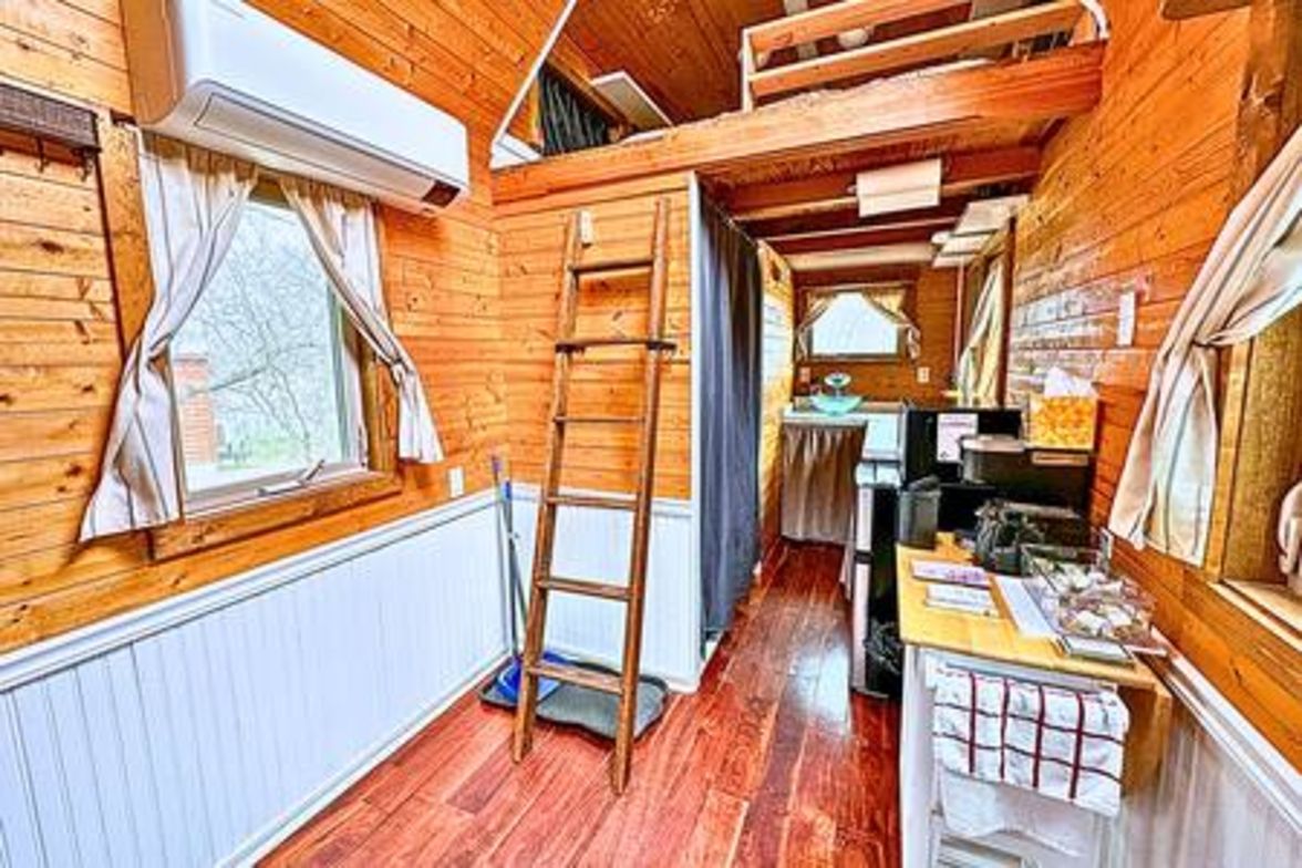 Een smal houten cabinteinterieur met een ladder, een raam met gordijnen en een klein kitchenette.