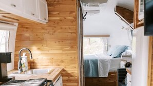 Cocina compacta para camper con paredes de madera, estufa, lavamanos y una pequeña zona de dormir con cama y ventana.