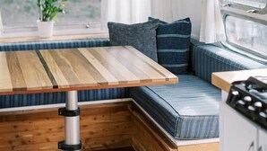Interior compacto de una caravana con una mesa de madera, asientos acolchados, una pequeña cocina y una ventana con cortinas.