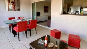 Un salon moderne avec une table à manger en verre, des chaises rouges, une table basse et un coin cuisine visible à travers une porte ouverte.