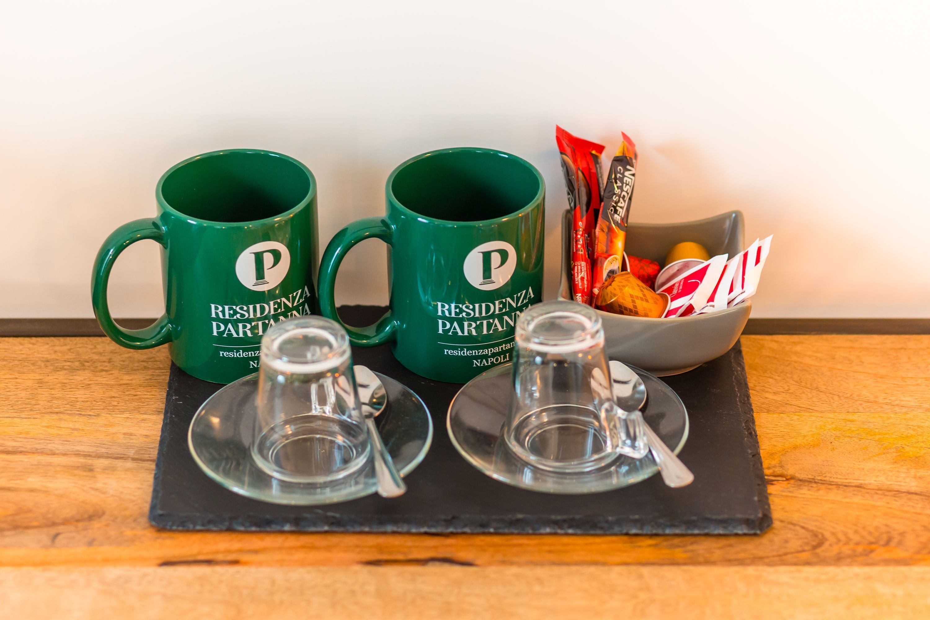 Two green mugs with "RESIDENZA PARTENZA" text, a bowl of snacks, and two empty glasses on a black tray.