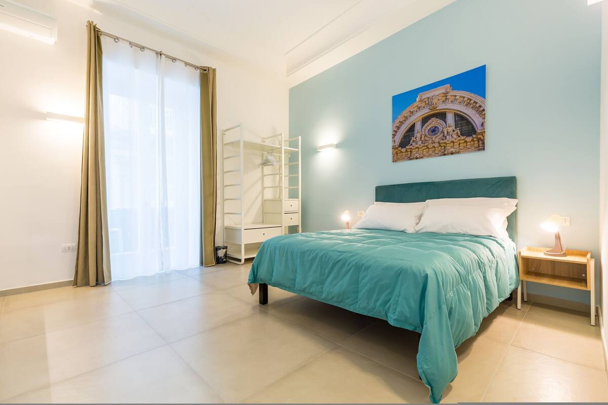 Ein Schlafzimmer mit einem Bett, einem Nachttisch, einer Wandablage, einem Fenster mit Vorhängen und einem Gemälde.